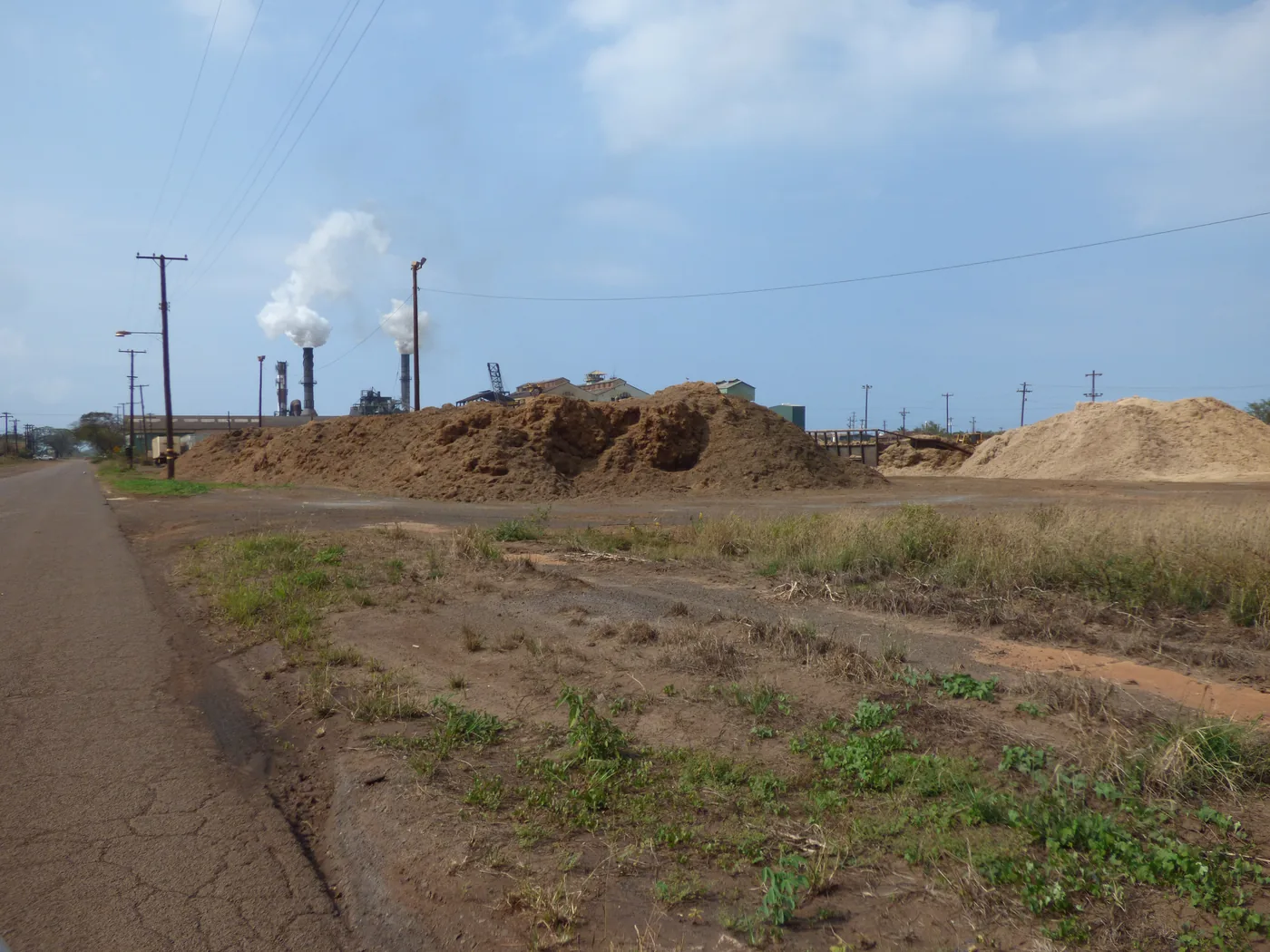 Large piles of bagasse beside a sugar mill with two stacks releasing steam