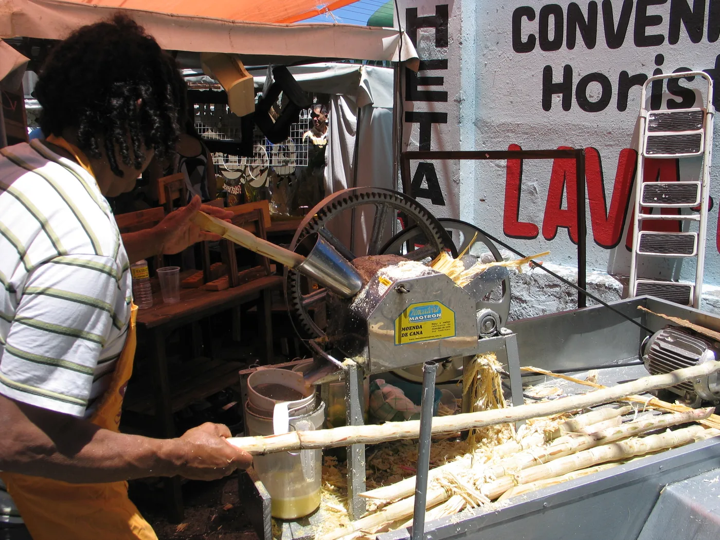 A vendor feeding cane stalks into a roller press at a stall, with crushed fibre and a cup of juice below